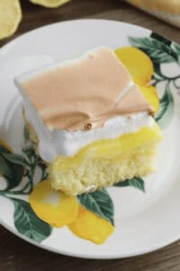 A slice of lemon poke cake on a decorative plate with yellow lemons and green leaves. The cake has a yellow filling and a browned meringue topping.