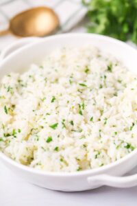 A white bowl filled with fluffy lime cilantro rice garnished with chopped herbs, with a gold spoon and green herbs blurred in the background.