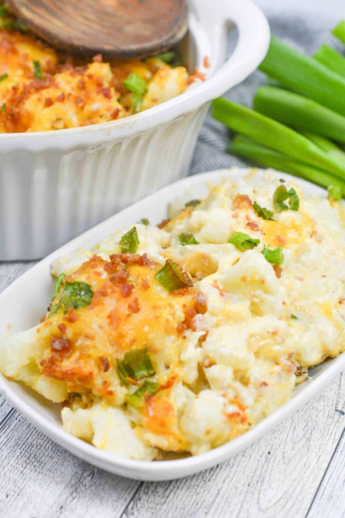 Savor the deliciousness of a Cheesy Cauliflower Casserole, topped with green onions and crispy bits. Served on a white plate, this dish is complemented by a white baking dish and fresh green onions in the background.