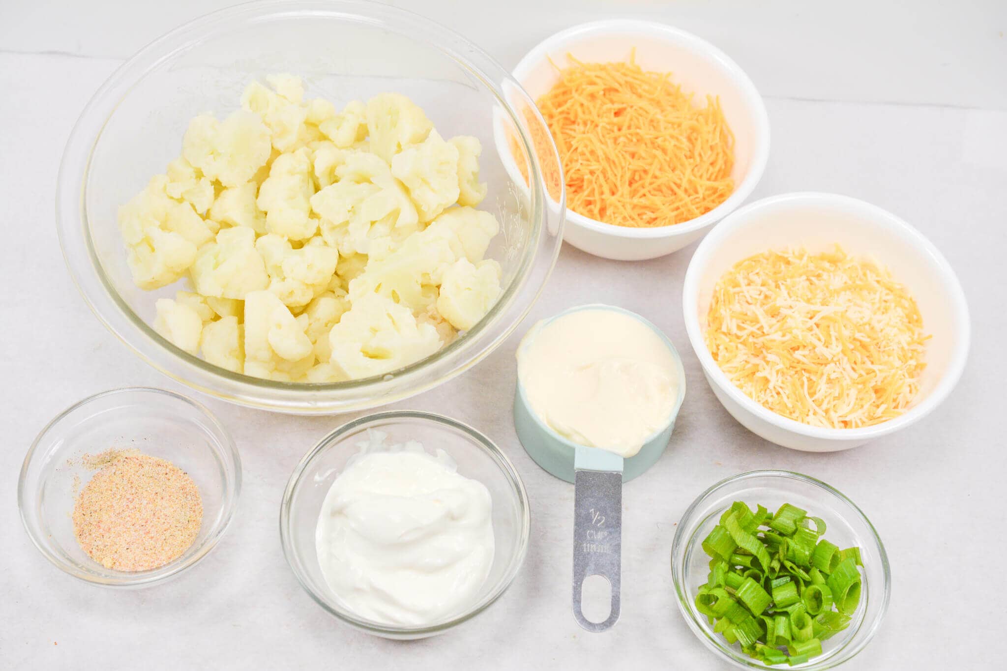 Ingredients for a cheesy cauliflower casserole include cauliflower, shredded cheese, yogurt, sour cream, green onions, and seasoning in glass bowls and a measuring cup on a white surface.