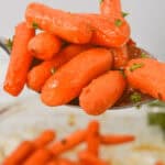 A spoonful of maple glazed carrots garnished with herbs, with more carrots visible in a dish below.