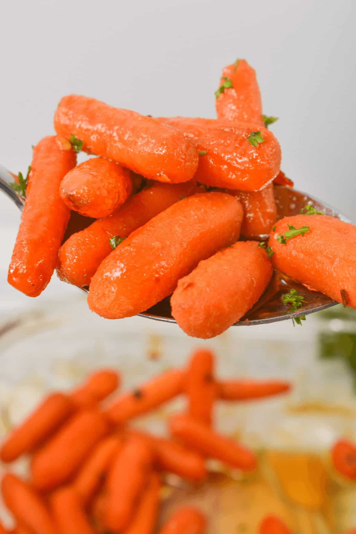 A spoonful of maple glazed carrots garnished with herbs, with more carrots visible in a dish below.