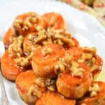 Maple Sweet Potatoes are sliced and topped with candied walnuts and a glossy glaze, all beautifully arranged on a white oval plate.