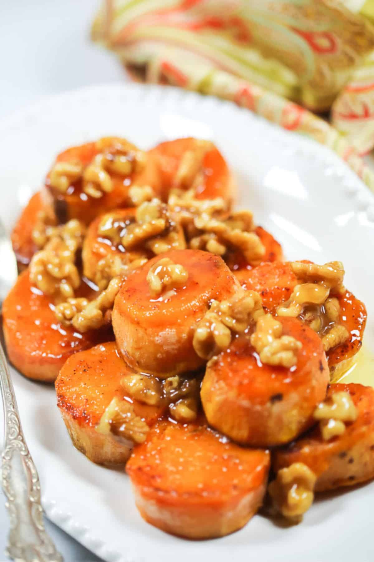 Maple Sweet Potatoes are sliced and topped with candied walnuts and a glossy glaze, all beautifully arranged on a white oval plate.