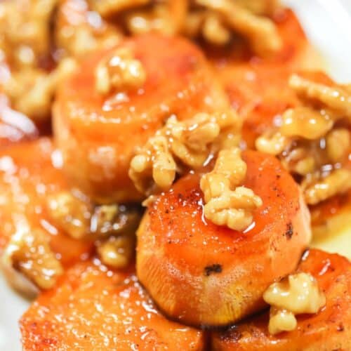 Sliced roasted Maple Sweet Potatoes topped with glazed walnuts on a white plate.