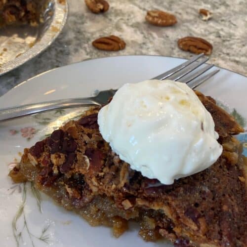 A slice of Maple Syrup Pecan Pie topped with vanilla ice cream rests on a plate, with a fork and scattered pecans on a gray surface in the background.