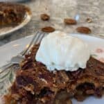 A slice of Maple Syrup Pecan Pie topped with a scoop of vanilla ice cream sits on a plate, with a fork and some pecans nearby.