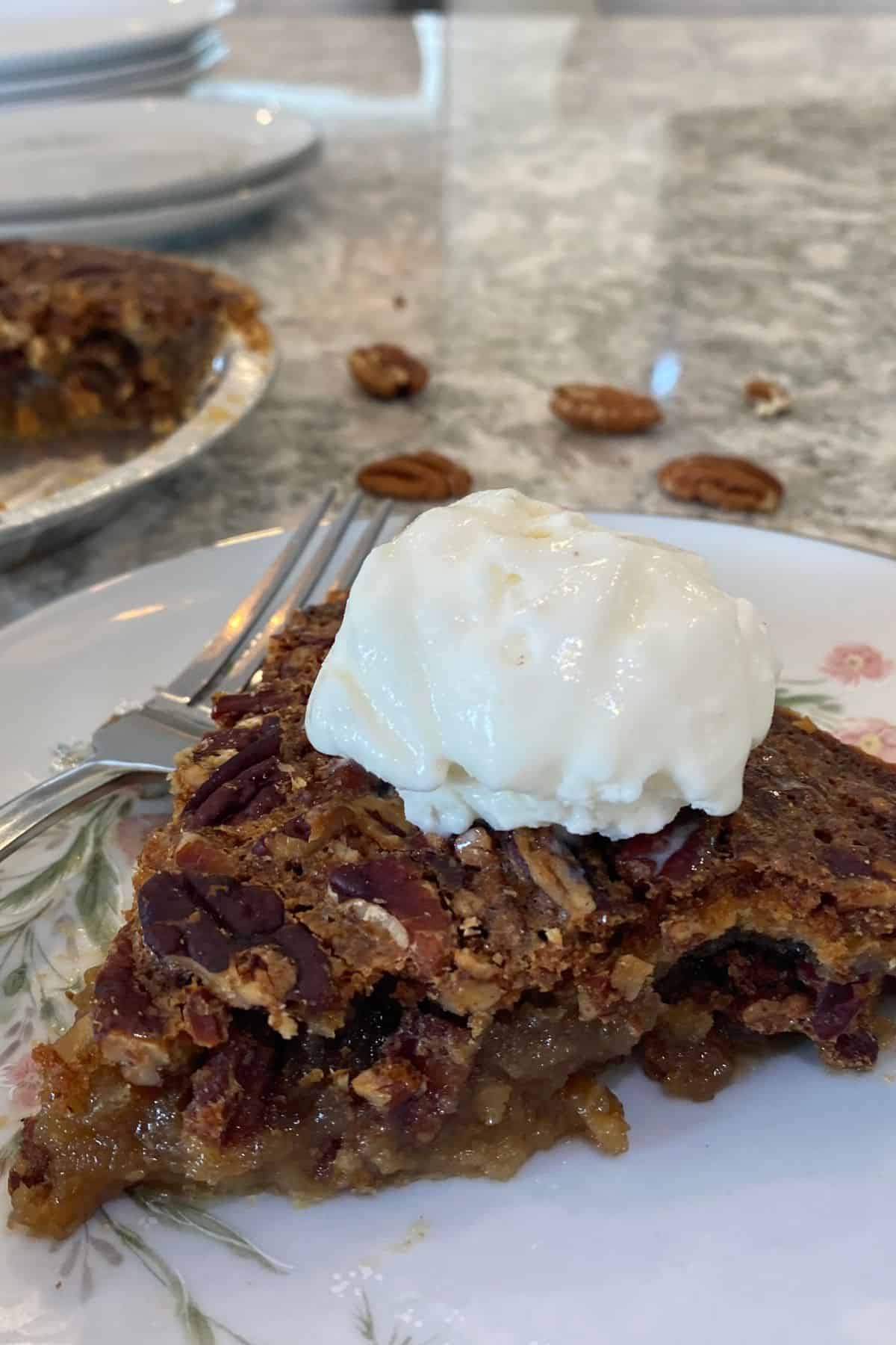 A slice of Maple Syrup Pecan Pie topped with a scoop of vanilla ice cream sits on a plate, with a fork and some pecans nearby.