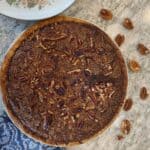 A dessert sits on a countertop next to a blue patterned towel, scattered pecan halves, and a floral-patterned plate.