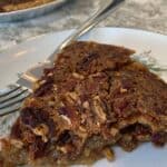 A slice of Maple Syrup Pecan Pie on a floral plate with a fork, showing gooey filling and pecan topping, with the rest of the pie in the background.