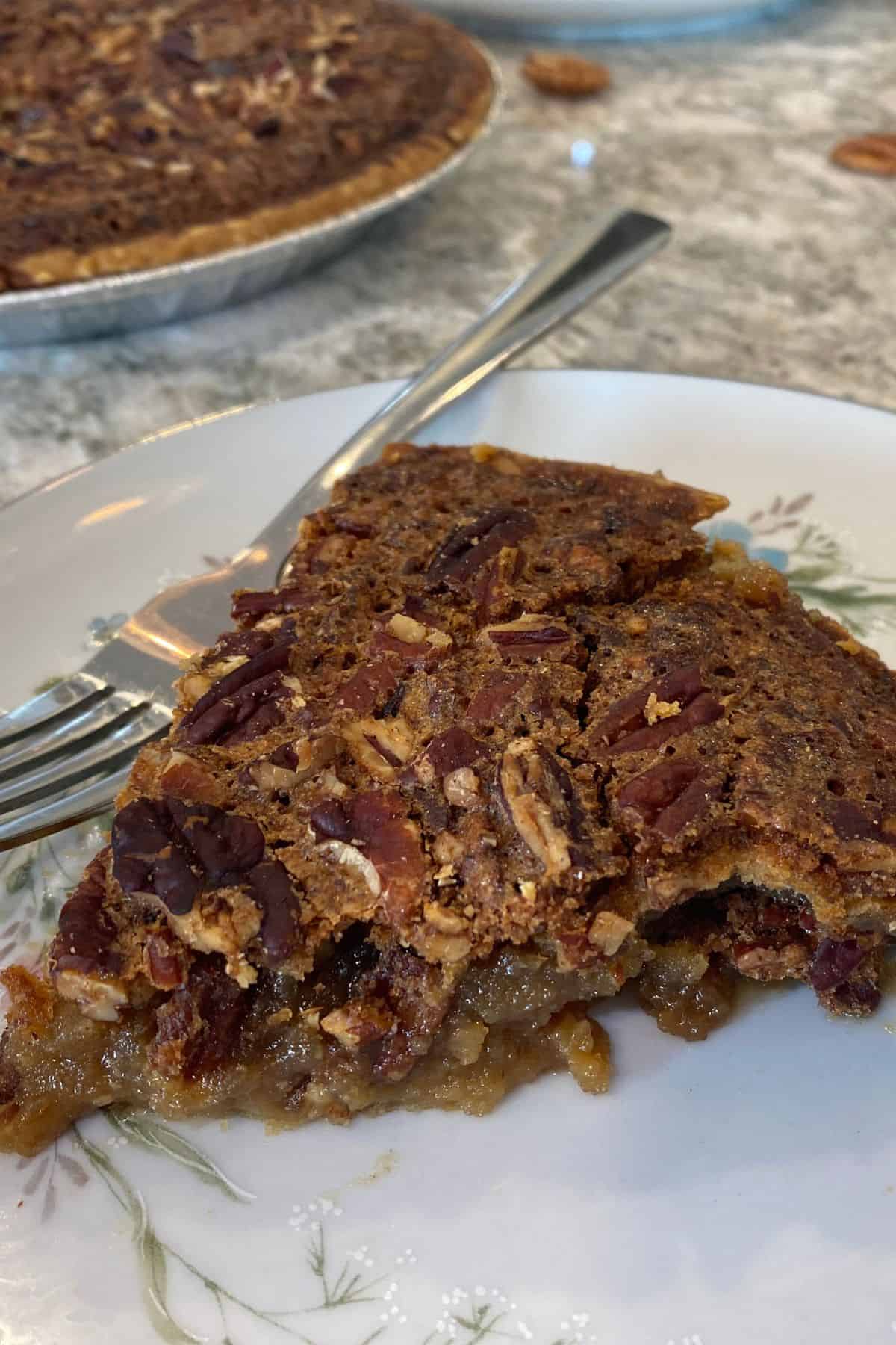 A slice of Maple Syrup Pecan Pie on a floral plate with a fork, showing gooey filling and pecan topping, with the rest of the pie in the background.