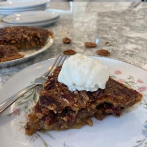 A slice of Maple Syrup Pecan Pie topped with a scoop of vanilla ice cream sits on a plate, with a fork beside it and more pie and pecans in the background.