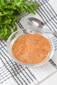 A bowl of meatloaf seasoning rests on a striped kitchen towel, surrounded by fresh parsley and a spoon.