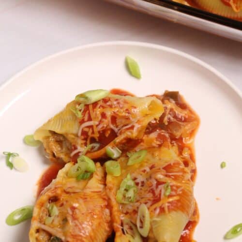 Three Mexican Stuffed Shells topped with tomato sauce, shredded cheese, and sliced green onions on a white plate, with more shells in a baking dish in the background.