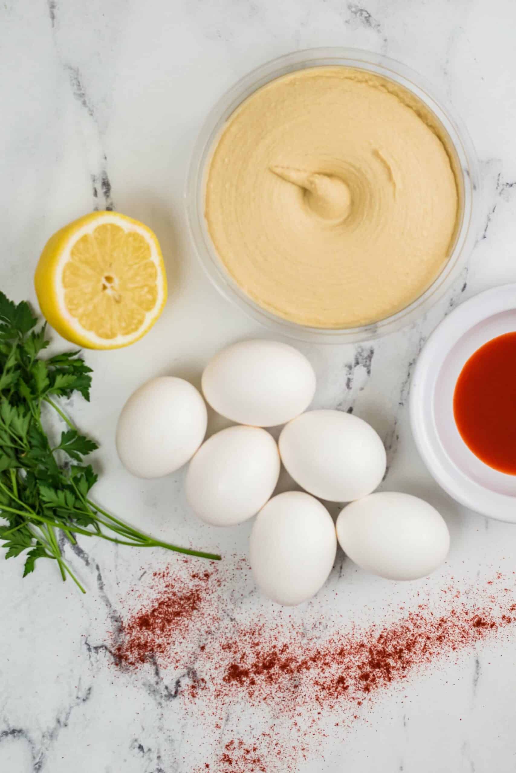 Eggs, paprika, hot sauce, parsley, lemon, and a bowl of creamy sauce, on a marble surface.