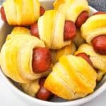 A bowl filled with several Mini Pigs in a Blanket, featuring mini sausages wrapped in golden, baked crescent roll dough.