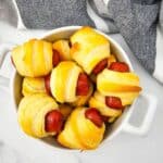 A white dish filled with several Mini Pigs in a Blanket, small sausages wrapped in golden-brown pastry, sits on a marble surface next to a striped cloth.