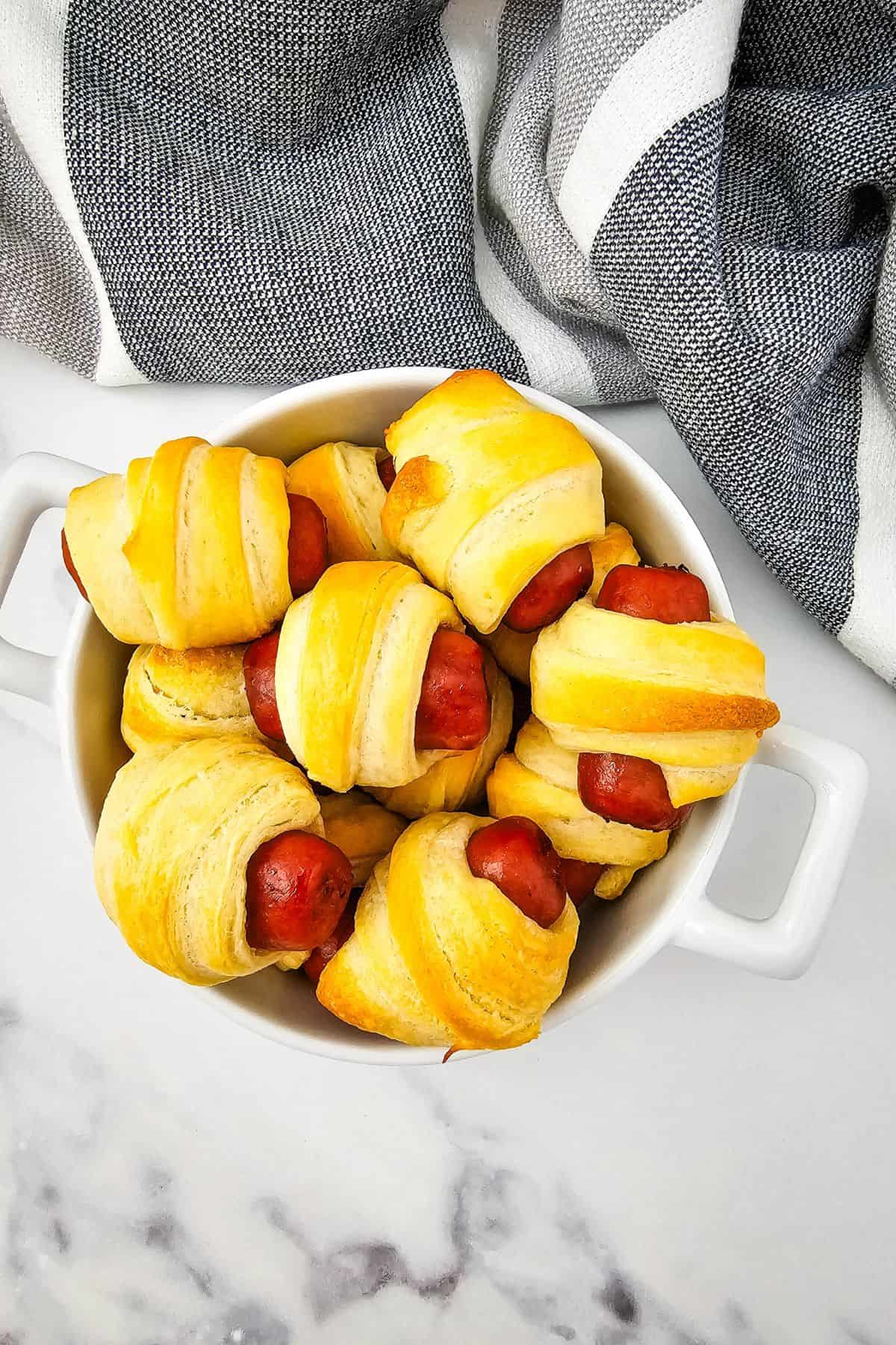A white dish filled with several Mini Pigs in a Blanket, small sausages wrapped in golden-brown pastry, sits on a marble surface next to a striped cloth.