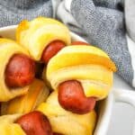 A plate of appetizers with mini sausages wrapped in golden-brown pastry, sits next to a folded gray and white cloth.