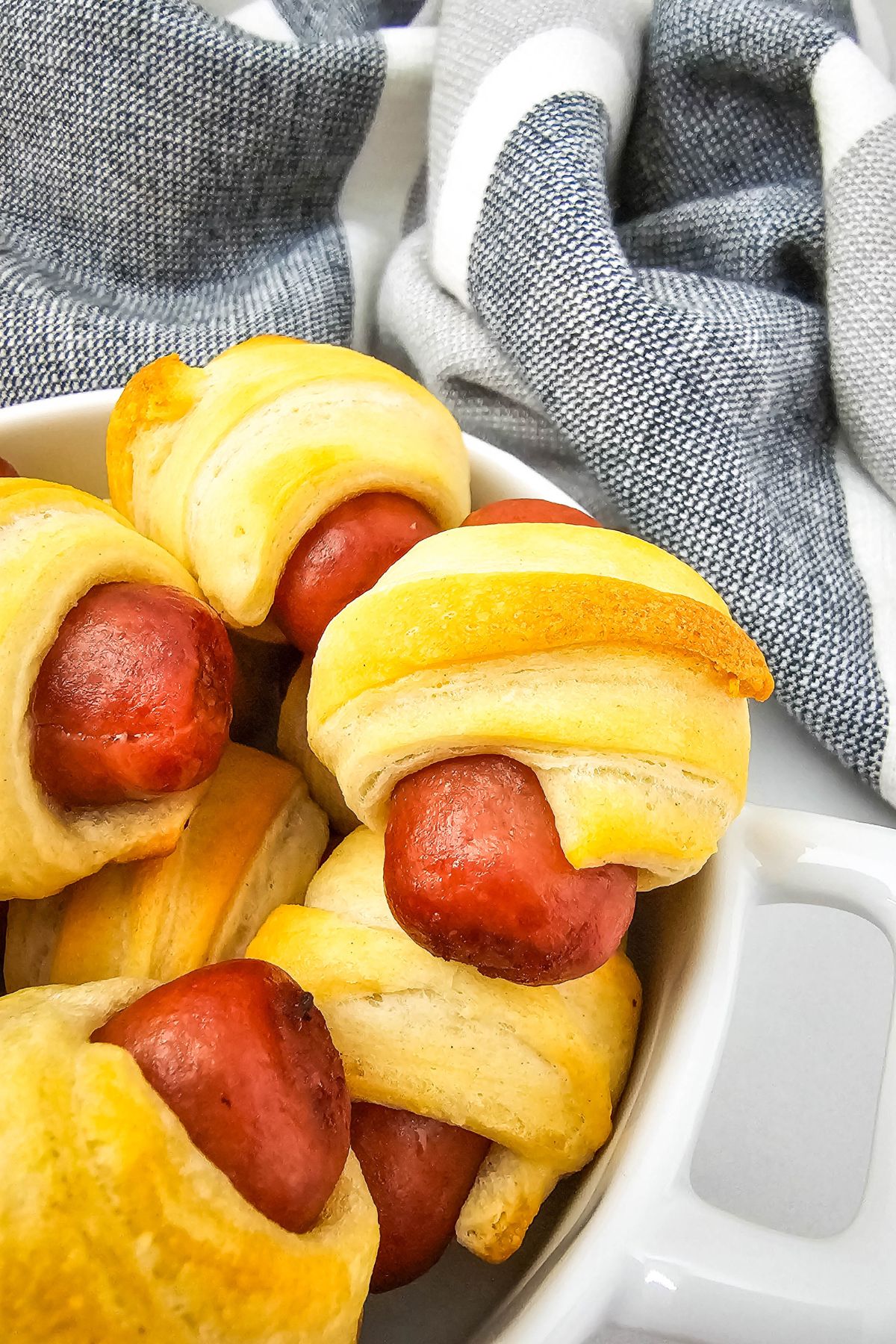 A plate of appetizers with mini sausages wrapped in golden-brown pastry, sits next to a folded gray and white cloth.