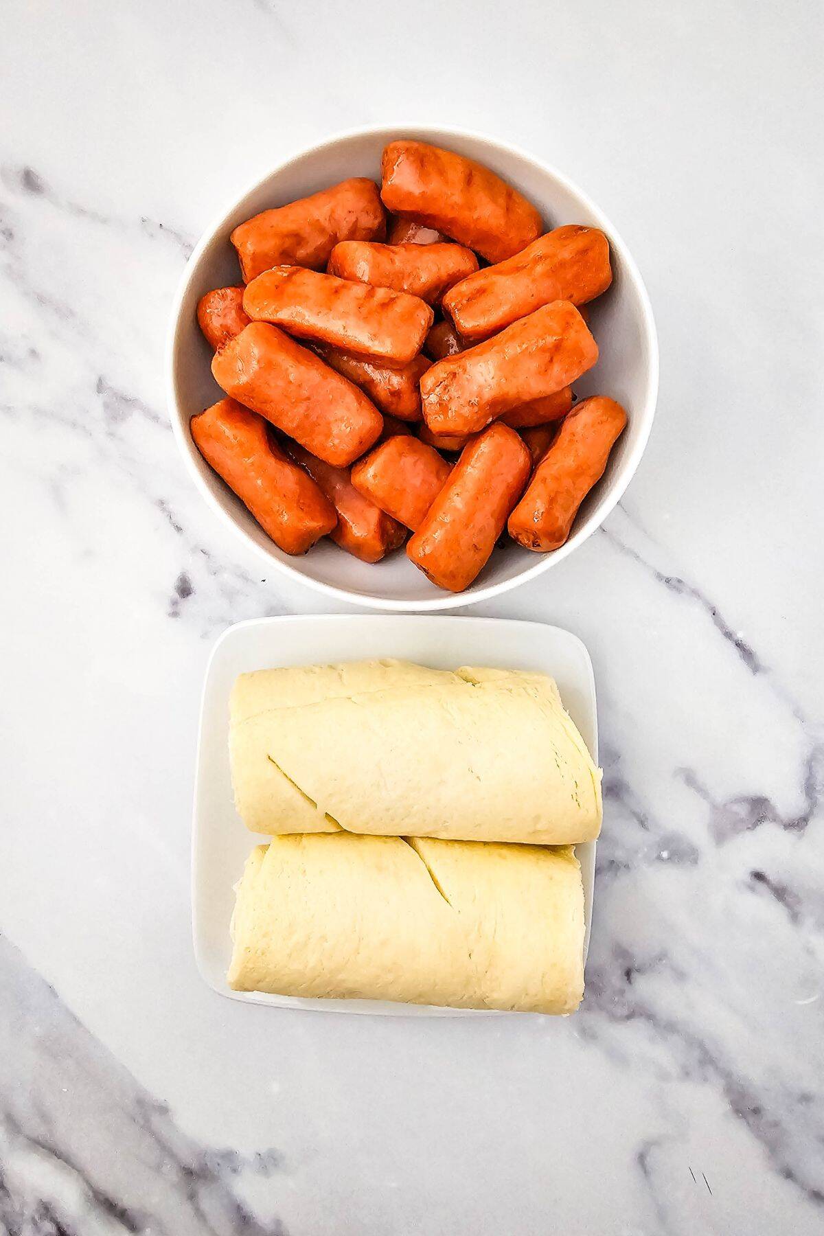 A bowl of small sausages sits above a plate with two pieces of rolled dough, perfect for making Mini Pigs in a Blanket, on a white marble surface.