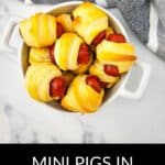 A white dish filled with Mini Pigs in a Blanket sits on a marble surface, next to a gray and white striped towel. Text below reads, "MINI PIGS IN A BLANKET.
