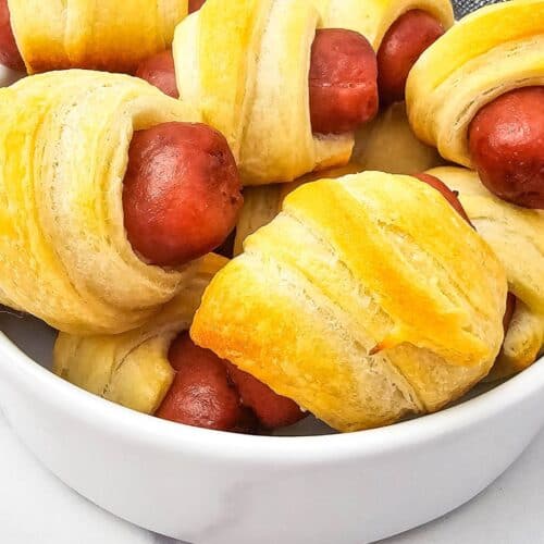 A white bowl filled with several Mini Pigs in a Blanket, featuring cocktail sausages wrapped in golden brown crescent roll dough.