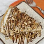 A slice of Butterfinger Pie with a graham cracker crust, topped with whipped cream and drizzles of chocolate and caramel sauce, on a square plate next to an orange napkin and fork.