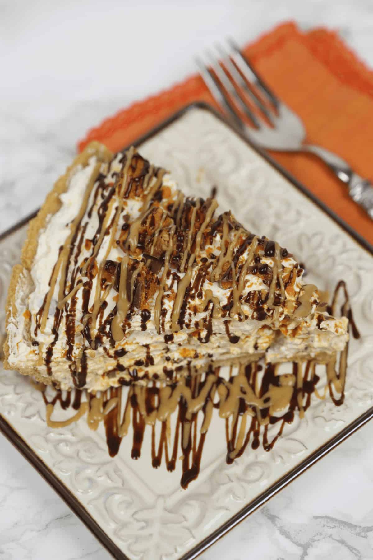 A slice of Butterfinger Pie with a graham cracker crust, topped with whipped cream and drizzles of chocolate and caramel sauce, on a square plate next to an orange napkin and fork.