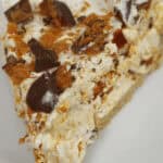 A slice of Butterfinger Pie with crumbled chocolate pieces and a creamy filling sits enticingly on a white plate.