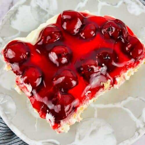 A slice of No Bake Cherry Cheesecake with a creamy layer sits on a white marbled plate, topped with cherries and placed next to a blue-striped cloth.