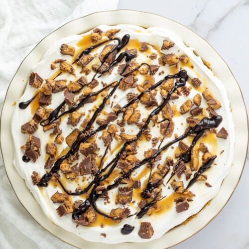 A round Chocolate and Peanut Butter Pie topped with whipped cream, chopped peanut butter cups, caramel sauce, and a chocolate drizzle on a white plate.