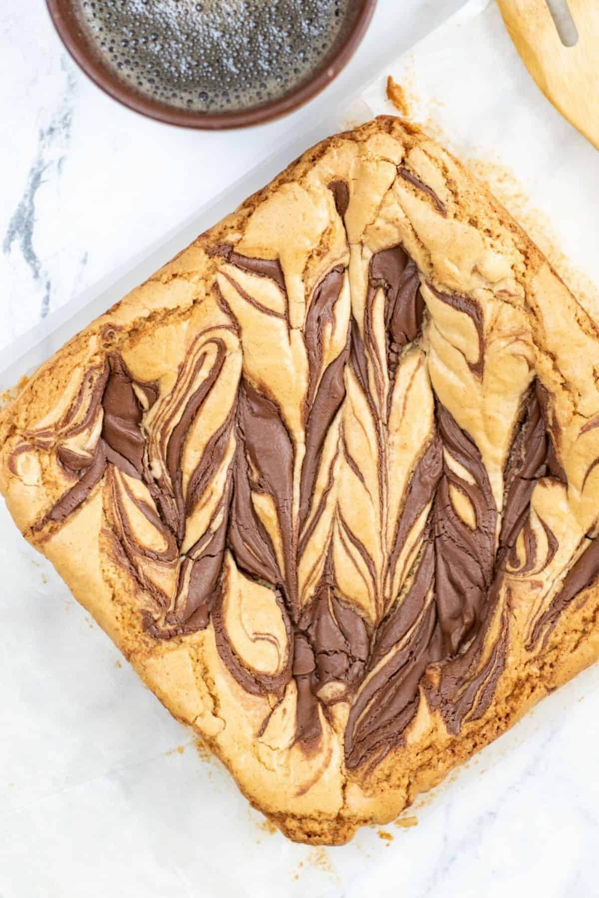 A tray of swirled chocolate and brown butter blondies sits on parchment paper beside a cup of black coffee on a marble surface.
