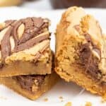 Two thick Brown Butter Blondies with a chocolate swirl on top are stacked on a white plate, with crumbs scattered around them.