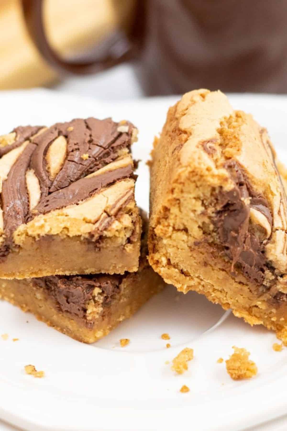 Two thick Brown Butter Blondies with a chocolate swirl on top are stacked on a white plate, with crumbs scattered around them.