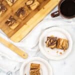 Sliced brown butter blondies on a wooden board, with two plates each holding two pieces, and a cup of black coffee on a marble surface.