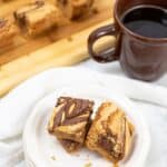 A plate with two squares next to a brown mug of black coffee; more bars are on a wooden board in the background.