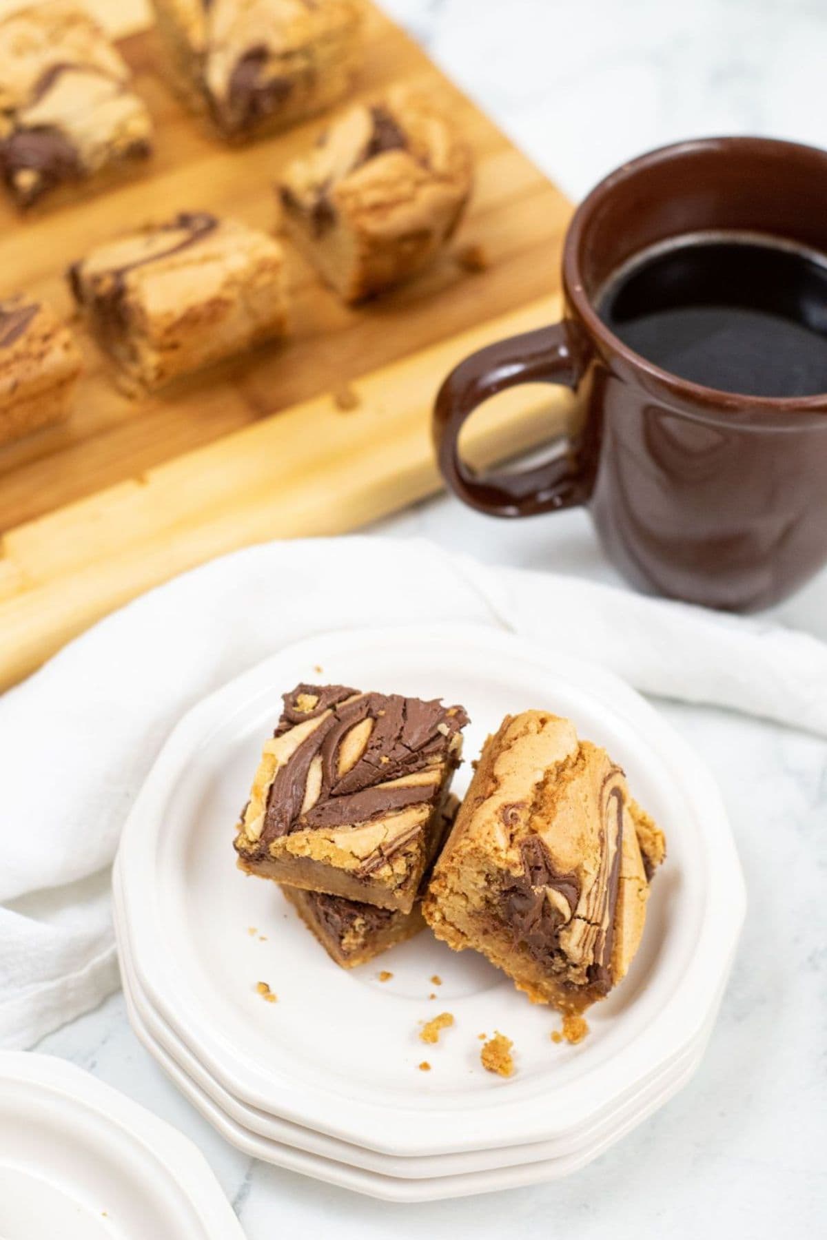 A plate with two squares next to a brown mug of black coffee; more bars are on a wooden board in the background.