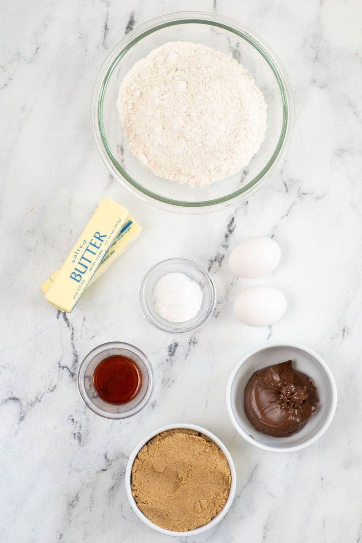 Baking ingredients on a marble surface: flour, butter, baking powder, eggs, vanilla extract, brown sugar, and chocolate spread—all ready to make delicious treats.
