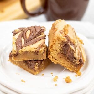 Two Brown Butter Blondies with peanut butter and chocolate swirls are stacked on a white plate, crumbs scattered around, with a brown cup in the background.