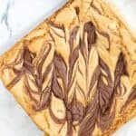 A square Brown Butter Blondie topped with swirled chocolate sits on parchment paper next to a cup of coffee on a marble surface.