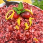 A bowl of Ocean Spray Cranberry Relish Recipe is garnished with lemon peel, whole cranberries, and a sprig of mint, with lemon halves in the background.