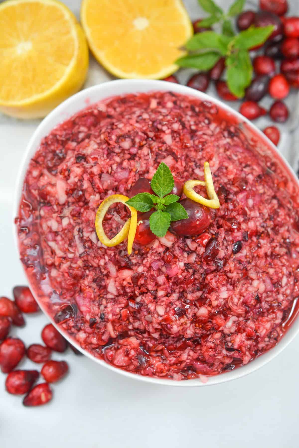 A bowl of Ocean Spray Cranberry Relish Recipe is garnished with mint, lemon peel, and cranberries, with orange halves and fresh cranberries in the background.
