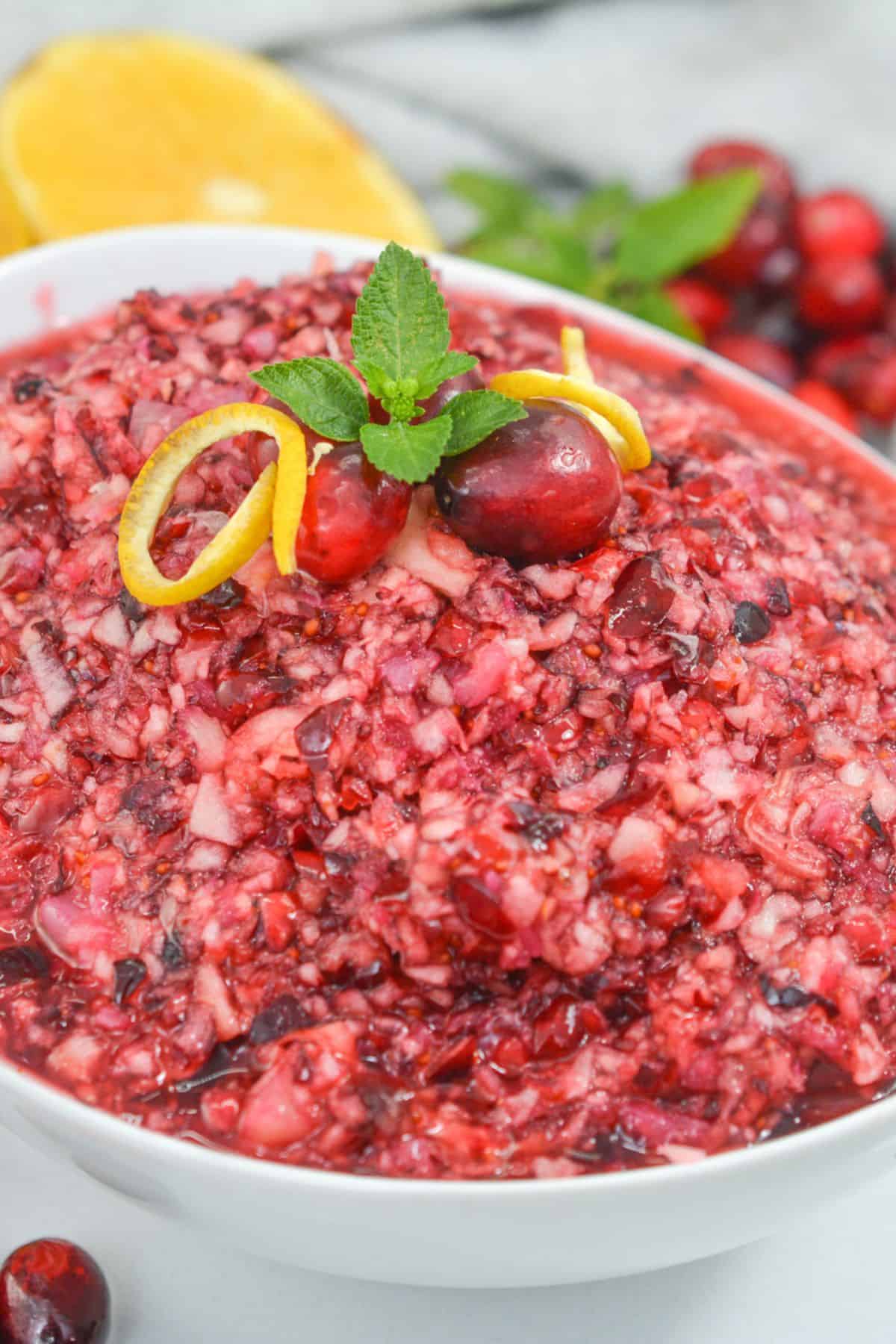 A bowl of the Recipe topped with whole fruit, a lemon twist, and a mint sprig, with a sliced lemon in the background.