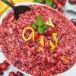 A bowl of Ocean Spray Cranberry Relish Recipe topped with mint and lemon zest, surrounded by fresh cranberries and halved lemons, with a spoon in the bowl.