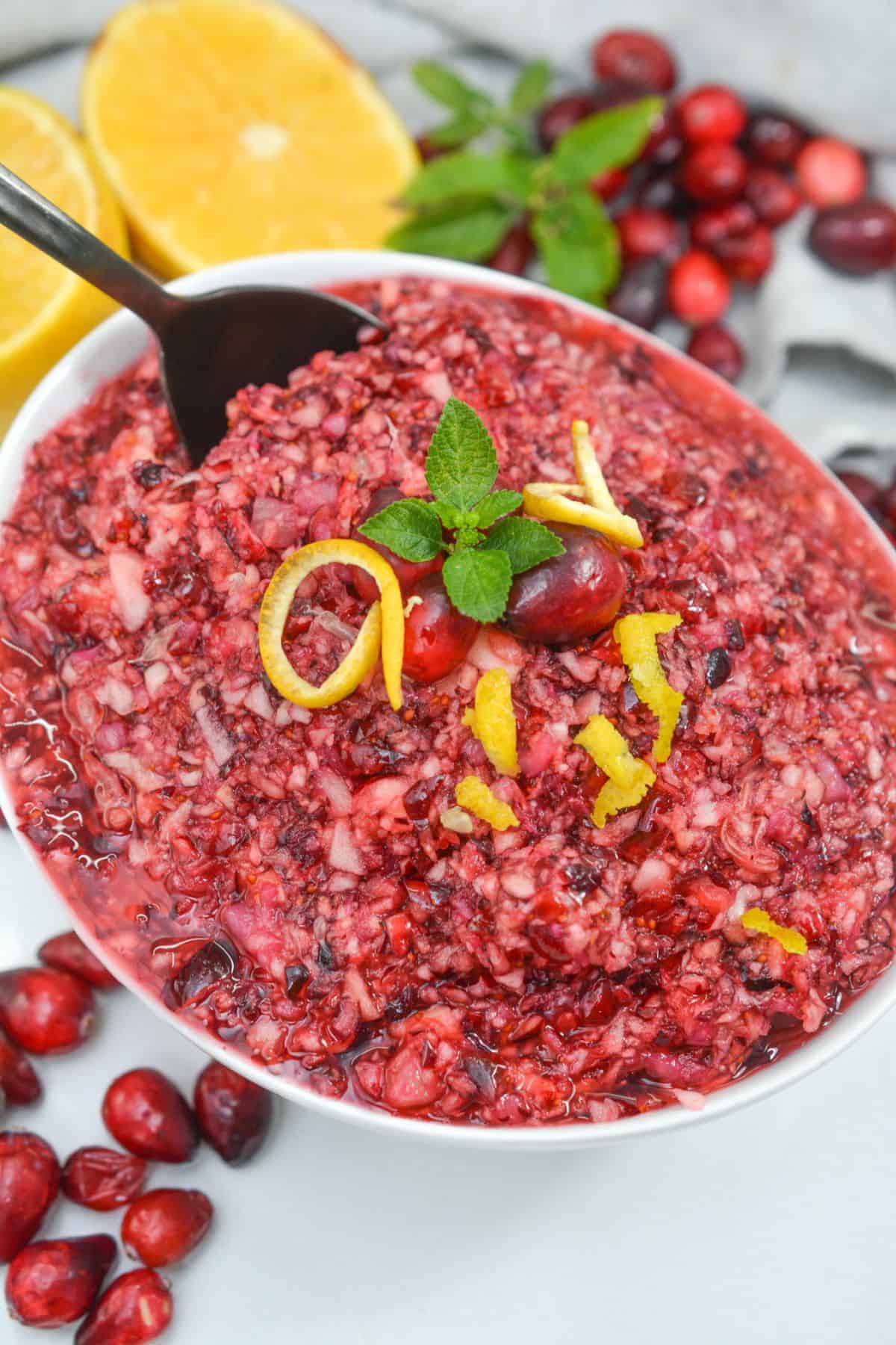 A bowl of Ocean Spray Cranberry Relish Recipe topped with mint and lemon zest, surrounded by fresh cranberries and halved lemons, with a spoon in the bowl.