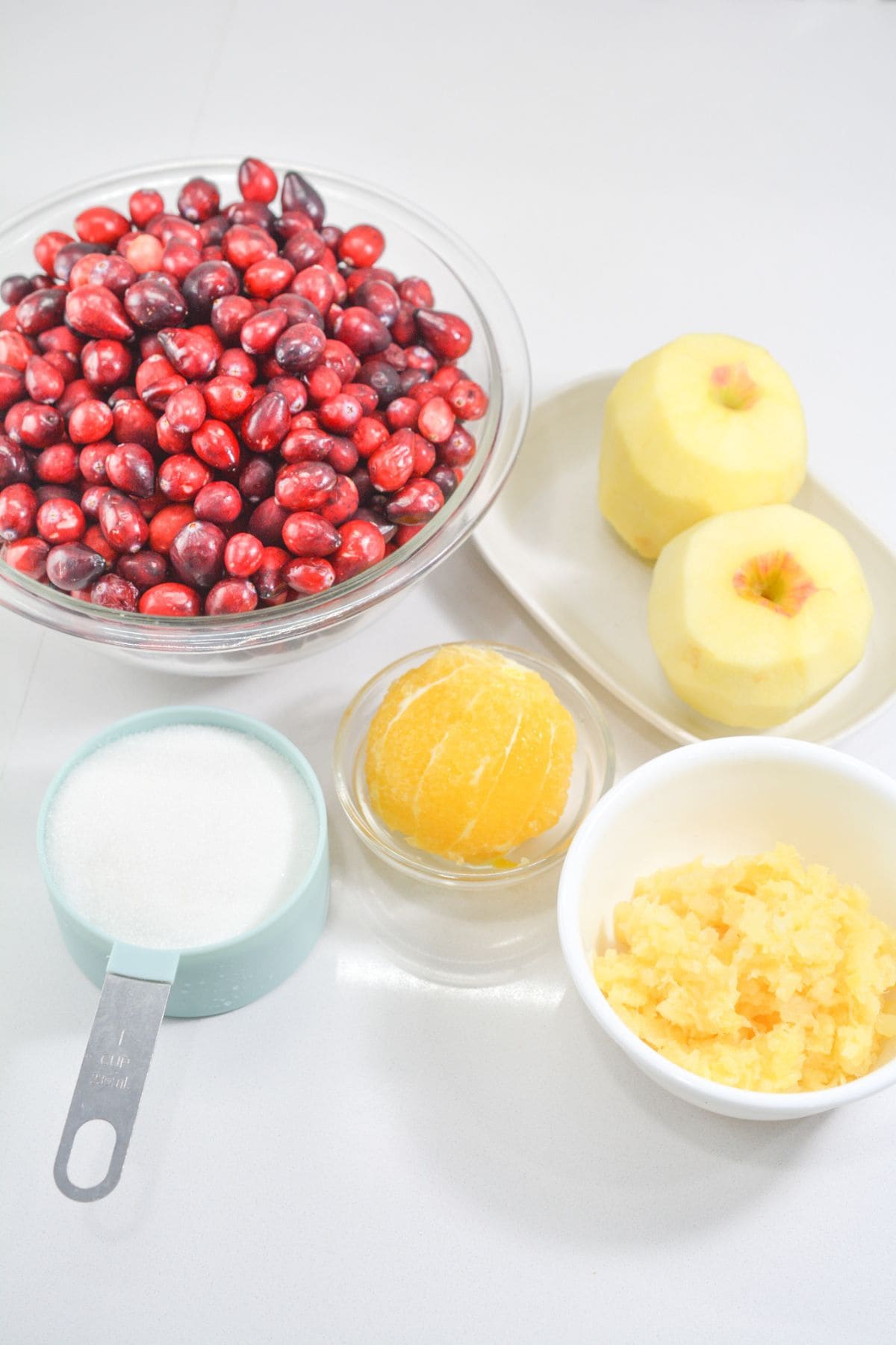 A bowl of cranberries, two peeled apples, a peeled orange, a measuring cup of sugar, and chopped ginger are arranged on a white surface—perfect ingredients for an Ocean Spray Cranberry Relish Recipe.