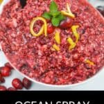 A bowl of Ocean Spray Cranberry Relish Recipe is garnished with cranberries, lemon peel, and mint, with a spoon in the bowl and fresh cranberries scattered nearby.