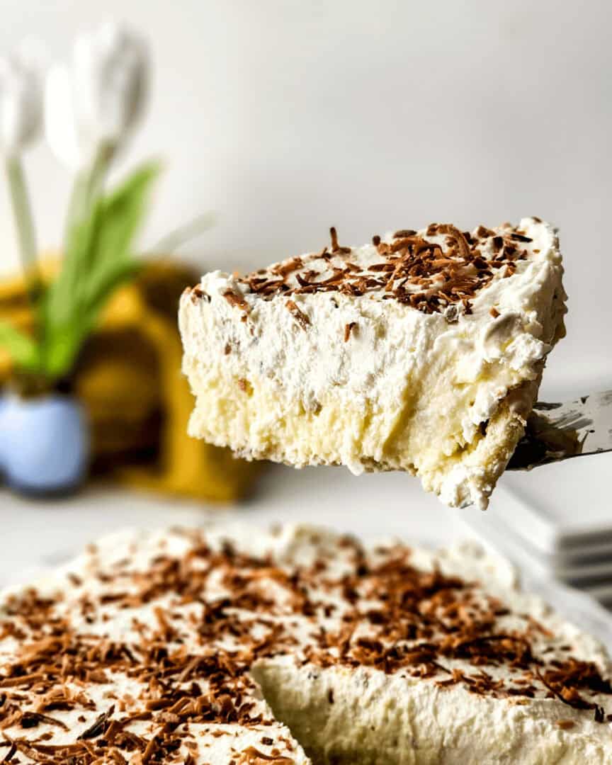 A slice of creamy old-fashioned coconut cream pie topped with chocolate shavings, with the remainder of the pie in the background.