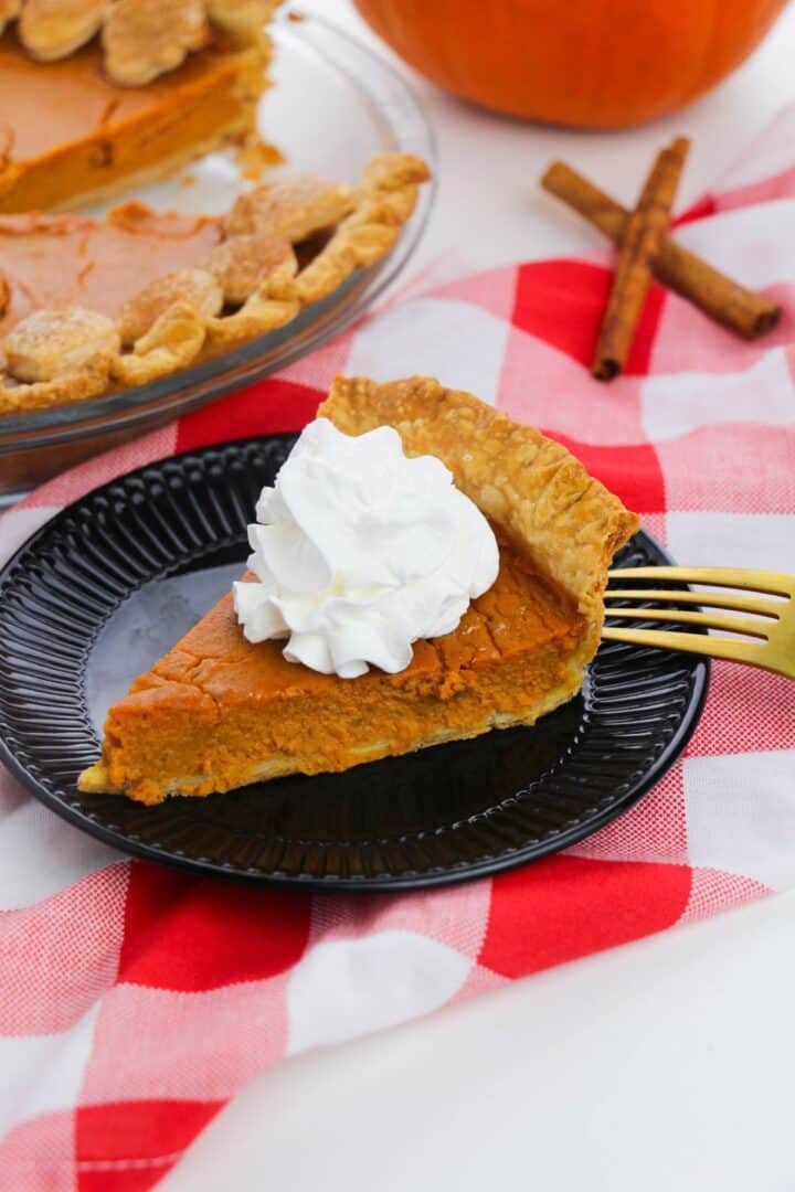 A slice of Old Fashioned Pumpkin Pie with whipped cream on a black plate, set on a red and white checkered cloth, with a gold fork and cinnamon sticks nearby.